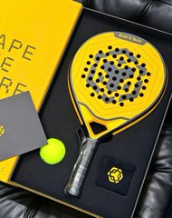 A black and yellow padel racket with holes displayed in a black gift box alongside a yellow ball.