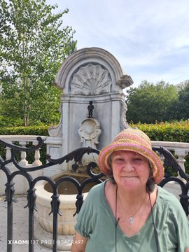 A smiling older woman wearing a colorful crocheted hat and a green shirt poses for a photo in front of an ornate stone fountain, taken with a Xiaomi 12 Pro.