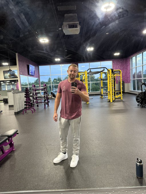 A man takes a mirror selfie inside a gym with purple accents and workout equipment.