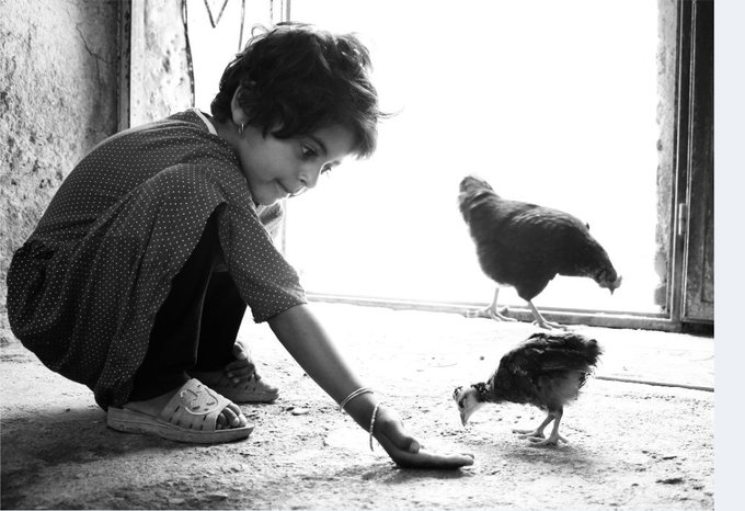 Black and white photograph of a young child squatting down to feed or interact with two chickens in a doorway.