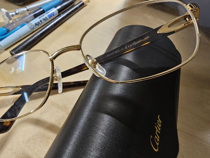A close-up shot of a pair of gold-rimmed Cartier eyeglasses resting on their black leather case, alongside a few pens on a wooden surface.