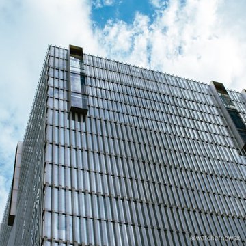 A low-angle shot of a modern, multi-story building with a glass facade against a partly cloudy blue sky, with a faint watermark '@ouchenihowa' in the corner.