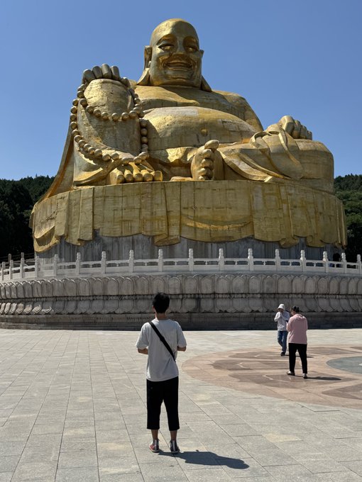 A young man stands before a massive golden Buddha statue at an outdoor temple plaza, with two other visitors nearby, as he enjoys a walk with his son on a sunny day while engaging with the #STEPN move-to-earn fitness app.