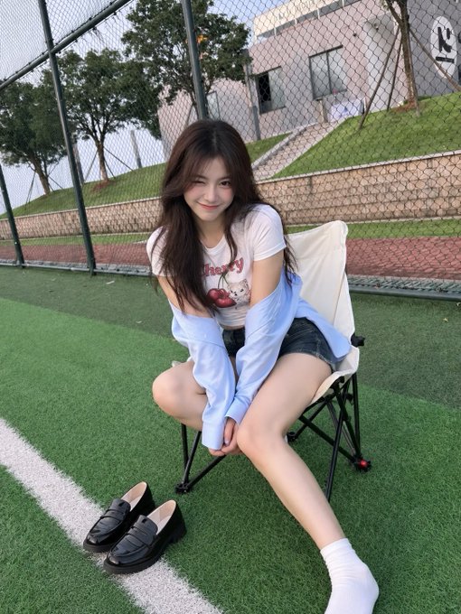 A young woman with long brown hair sits on a folding chair on a sports field, smiling at the camera while wearing a white t-shirt, black shorts, and a light blue jacket draped over her arms.