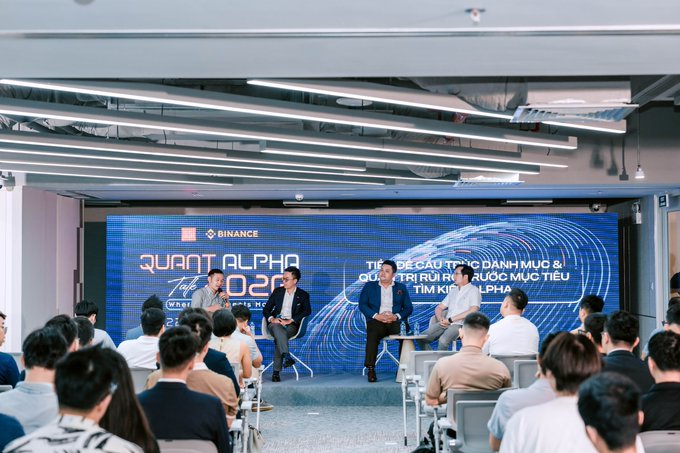 A panel discussion event for 'QUANT ALPHA 2026' hosted by Binance, featuring speakers on stage discussing portfolio creation and pursuing investment targets, viewed from the audience seated in the foreground.