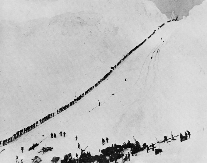 A black and white historical photograph showing a long line of people stretched diagonally across a snow-covered mountain slope, with a group of sled dogs and handlers gathered in the lower right area.