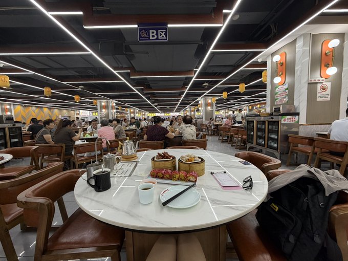 Interior of a bustling Chinese restaurant with section B marked, featuring round marble tables, dim sum steamers, and many diners under bright linear lighting.