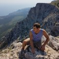 Photo of a woman in a purple tank top sitting on a rocky mountain peak, posing for the camera with mountainous terrain visible behind her.