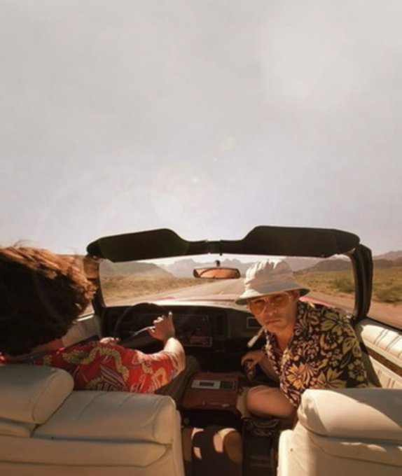 Two people riding in a convertible through a desert landscape, with the driver wearing a bucket hat and sunglasses glancing back, evoking a Fear and Loathing in Las Vegas aesthetic.
