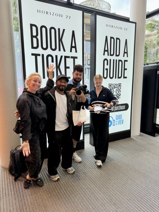 Four people pose enthusiastically in front of two Horizon 22 displays reading "BOOK A TICKET" and "ADD A GUIDE," with one person holding a camera, suggesting they are excited visitors or content creators sharing their Breakpoint experience.