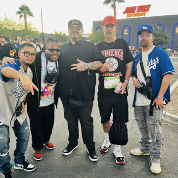 A group photo of six men posing outdoors, with the central figure wearing a 'Hoonigan' racing jacket and others in streetwear, taken at what appears to be a motorsport event.