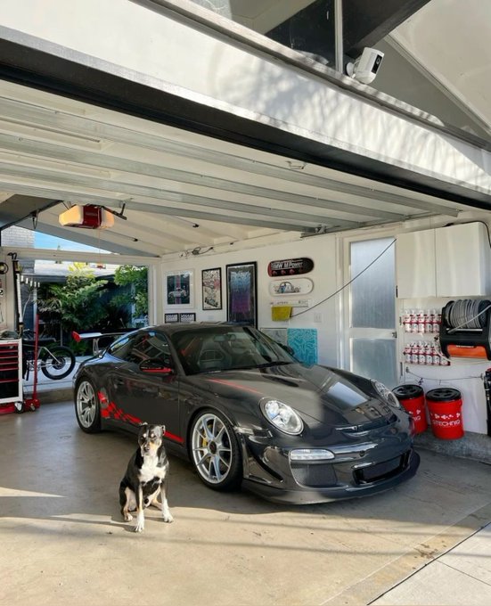 A black Porsche parked in a garage with a dog sitting in front of it, surrounded by tools and storage.