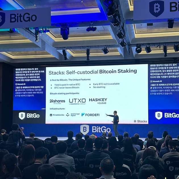 A presenter stands on stage at the BitGo event, pointing toward a large screen displaying "Stacks: Self-custodial Bitcoin Staking" featuring partners 21shares, HashKey Cloud, Anchorage Digital, and Fireblocks, illustrating the institutional participation—including 21shares—referenced in the post.