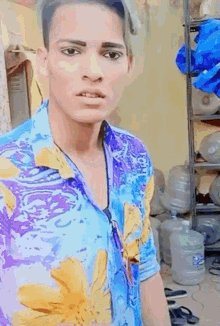 The image features a man wearing a blue shirt with yellow flowers printed on it. He has short hair and is looking directly at the camera. The background includes a shelf holding various items, such as a water bottle and a pair of scissors. There are also two other people in the scene, one standing behind the man and another person partially visible to his right side. The image contains text that reads "Okay $OFC is number 1."