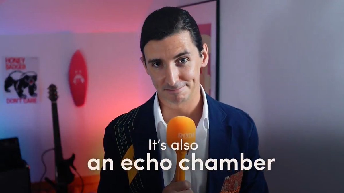 A man in a dark suit and white shirt holds an orange microphone with the text 'It's also an echo chamber' overlaid at the bottom of the frame.