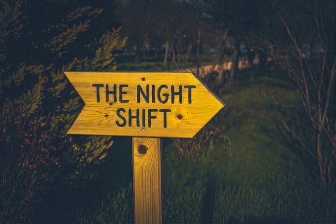 A weathered wooden signpost points right with the text "THE NIGHT SHIFT" painted in bold black letters, set against a dark, moody outdoor backdrop of grass and bare trees. The sign visually echoes the post's question "Who's up?"—inviting the nocturnal crowd, night owls, and after-hours workers to follow the arrow into the late-night hours.