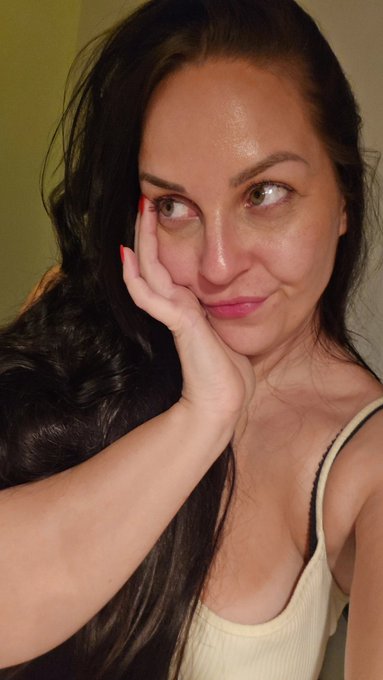 A close-up selfie of a woman with long dark wavy hair and red nail polish, resting her cheek on her hand with a slightly playful expression.