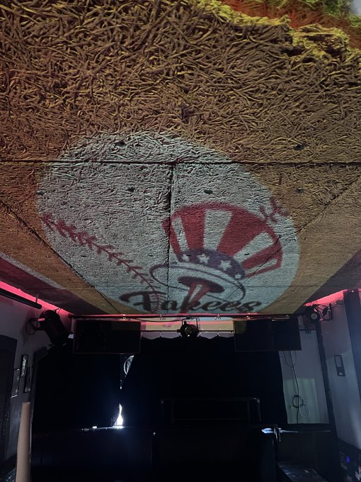 The image shows a large room with a ceiling that has a baseball logo painted on it, giving the impression of a baseball stadium or arena. The room also features a stage area where a DJ can be seen performing for an audience. A person is visible in the background, possibly enjoying the event. The post includes information about a new immersive experience called "The worlds first immersive Pepe" and mentions that it was brought to you by @BTCPrague and @FakeDjPepe.