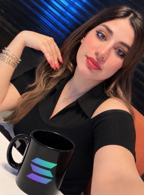 A selfie of a woman with long brown hair and red lipstick wearing a black top and crystal bracelet, posing next to a black mug featuring a holographic-style Solana logo.
