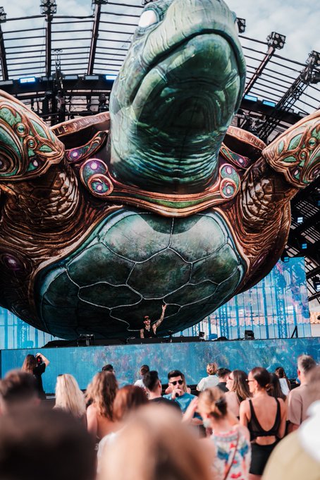 A DJ performs beneath a massive sea turtle stage sculpture at what appears to be an electronic music festival, with "gm future millionaires" echoing the upbeat, dream-chasing energy of the vibrant scene.