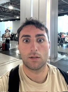 A man in a beige t-shirt displays a shocked, wide-eyed expression while taking a selfie in what appears to be an airport or convention center, likely capturing his disbelief after being eliminated from a live trading tournament.