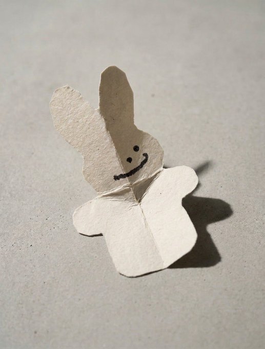 A simple paper origami figure with long rabbit-like ears sits on a plain surface, featuring a hand-drawn black smiling face.
