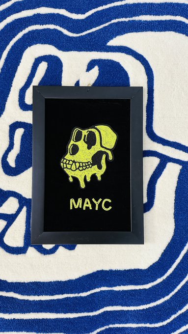The image features a black frame with a yellow cartoon depiction of a skull, which appears to be melting or dripping. The frame has the word "mayc" written on it, indicating that this artwork might be related to the "mutant ape yacht club". The background is blue and white striped, creating an interesting contrast against the black frame and the yellow skull.