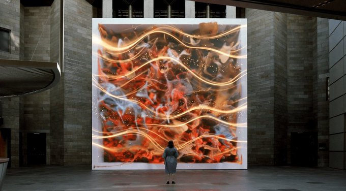 The image features an indoor space with a large screen displaying a vibrant display of orange and red lines against a gray background. A woman stands in front of this captivating artwork, seemingly mesmerized by its beauty. The scene also includes a brick wall and a window, adding depth and context to the setting. The post about @quipnetwork discusses the concept of a decentralized computing marketplace that combines classical and quantum resources, which could be related to the artistic display or even the technology behind it.
