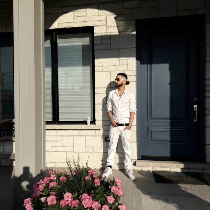 A man in an all-white outfit stands confidently against a stone wall between a window and a dark blue front door, projecting an image of casual wealth. The "POV: You hold $ANSEM" caption pairs with the scene to humorously suggest the lifestyle associated with holding the cryptocurrency, where modest, everyday luxury becomes the backdrop for crypto gains.