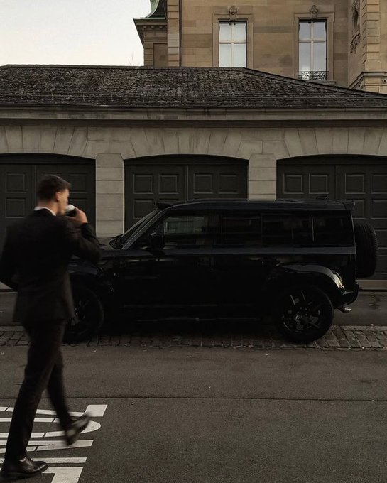 A photograph of a man in a black suit walking past a parked black Land Rover Defender on a European street with stone buildings and garage doors.