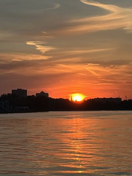 A sunset scene over a body of water with the orange sun low on the horizon, reflecting on the water, with silhouetted buildings and clouds in the sky.