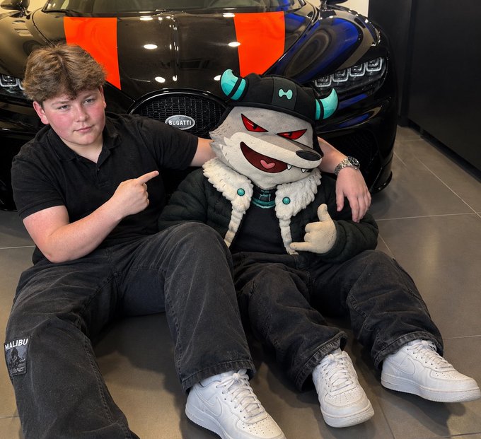 A young man sits beside a cartoonish mascot character in front of a black Bugatti sports car, both giving a thumbs up, while the man points at the "MALIBU" text on his jeans. The Bugatti and flashy lifestyle pairing suggests the post is hinting at wealth or a sponsorship deal, fitting the "Is this a sign?" caption.