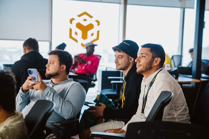 Three men sitting in an office looking attentively at something, with a yellow Binance logo overlay in the background.