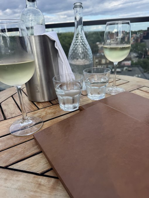 A leather-bound menu rests on the patio next to two glasses of white wine and a chilled water bottle, with a city skyline visible in the background—setting the scene for a well-earned "Happy Hour."