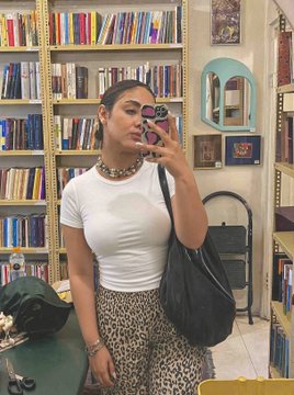 A mirror selfie of a woman in a white t-shirt and leopard print pants holding a camera in what appears to be a bookstore, with bookshelves visible in the background.