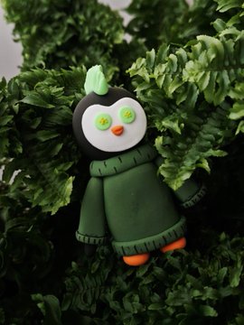 A small handmade penguin-like figurine with a green crest, yellow button eyes, an orange beak and feet, and a dark green coat sits among lush foliage.