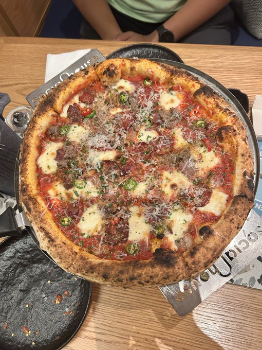 A freshly baked Neapolitan-style pizza topped with tomato sauce, melted mozzarella, pepperoni, jalapeños, and grated cheese sits on a black pan at a restaurant table, with what appears to be a "Social Order" or similar branded item nearby. The $25 price tag mentioned in the post seems reasonable for this generously topped, wood-fired pizza, making it a solid "smash" in my book.