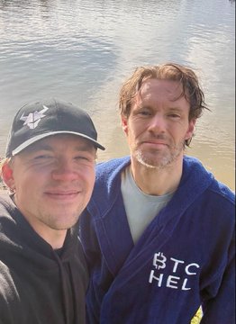 Two men pose for a selfie near a body of water. The man on the left wears a dark cap and hoodie, while the man on the right wears a blue bathrobe over a gray shirt.