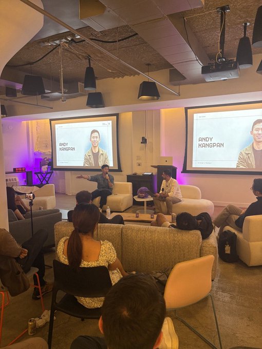 A photo of an indoor event or talk show with two people seated on couches while a screen displays 'ANDY KANGPAN' and an audience watches from the foreground.