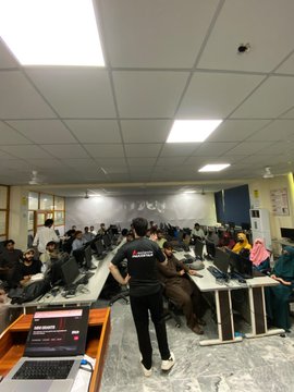 A crowded computer lab or office space shows many people seated at desks working on laptops, with one person standing in the center wearing a black shirt with 'Libris' written on the back.