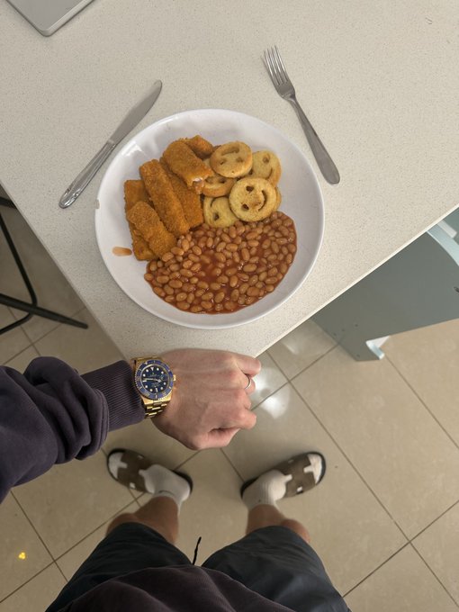 An overhead first-person view of a person standing at a white table wearing a two-tone gold and silver watch (resembling a Rolex), about to eat a meal of fried hash browns, smiley-face potato faces, and baked beans in a white bowl, with a knife and fork set on either side.