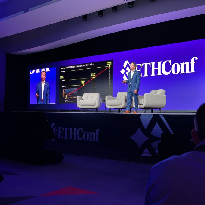 The image shows a man standing at a podium, giving a speech or presentation. The room has a stage set up with chairs arranged around it, and there are two screens displaying information related to the event. A person can be seen sitting on one of the chairs in the background. The text "@BitMNR is still bullish on $ETH @ethconf Thank you for your attention to this matter" suggests that the man speaking might have some connection or involvement with BitMNR, and he could be discussing the current state of cryptocurrency markets or other related topics.