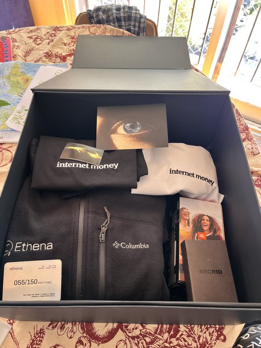 An open black PR gift box from Ethena displays a Columbia fleece jacket, internet money apparel, a Secrid cardholder, and a numbered edition card marked "055/150," reflecting the post's excitement about receiving a premium welcome gift from @ethena.