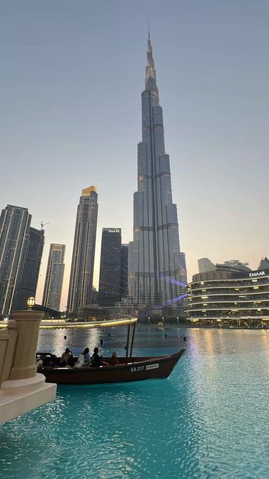 A traditional abra boat carrying passengers glides across the turquoise waters of the Burj Khalifa Lake in Dubai, with the towering Burj Khalifa dominating the skyline at dusk and the "EMAAR" branding visible on nearby buildings—fitting for a post simply tagged "GM" (Good Morning) showcasing an iconic Dubai sunrise view.
