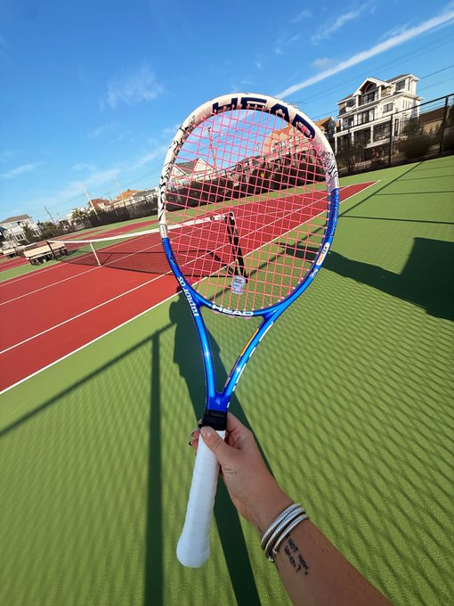 A hand holds a blue and white HEAD tennis racket on a tennis court, with houses and a blue sky visible in the background.