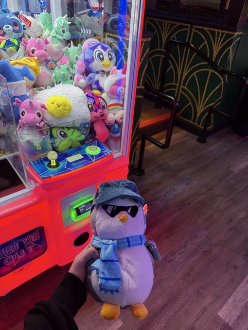 A hand holds a purple penguin plushie dressed in a denim hat, sunglasses, and blue scarf outside a brightly lit claw machine filled with colorful prizes. The machine's control panel reads "PRIZE OUT," celebrating the arcade win of the @pudgypenguins plush.