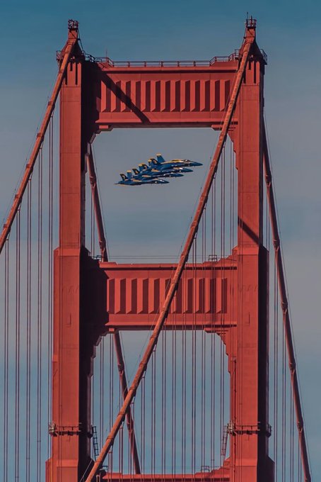 Four Blue Angels jets fly in a precise diamond formation through the iconic red towers of the Golden Gate Bridge against a clear blue sky.