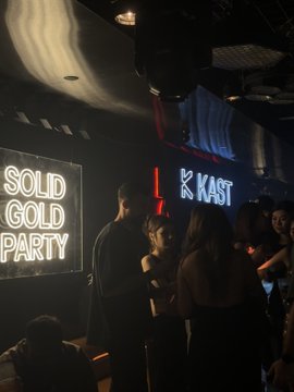 People mingle at a club party, with illuminated signs reading 'SOLID GOLD PARTY' and 'K KAST' visible in the background.