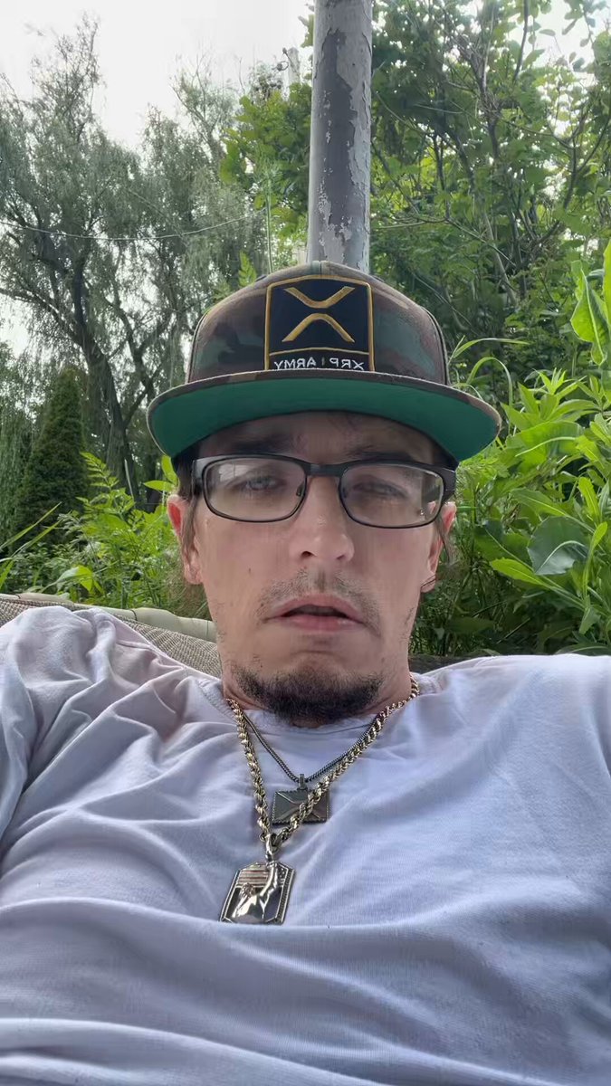 The image features a man wearing glasses, a white t-shirt with a gold chain necklace, and a black and yellow baseball cap. He appears to be sitting outdoors, possibly on a chair or bench, as he looks at something off camera. The caption of the image reads "I’m on that XRP", which is likely related to the post about RippleXRP.