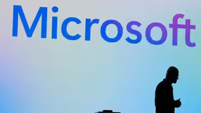 The Microsoft logo is displayed in blue on a light background, with the silhouette of a person walking by in the lower right corner. The image visually anchors the post's discussion of Microsoft's massive $190 billion infrastructure spending and GPU rationing challenges.